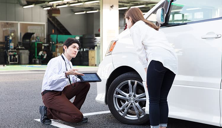 車の前でタブレット端末を用いて説明する整備士の男性や女性客|タイヤ館
